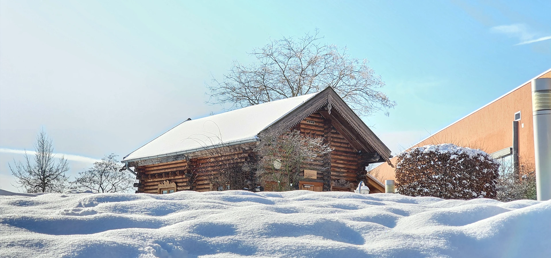 Inmitten des mit Schnee bedeckten Saunagartens steht unsere Banja. Umrandet von einem hellblauen Himmel und mit Schnee Akzenten versehenen Bäumen und Büschen.